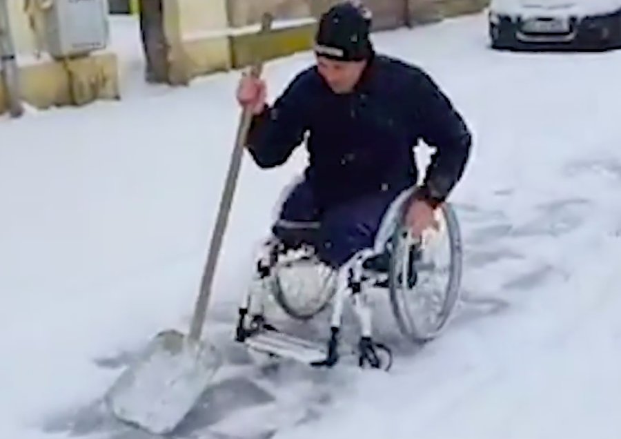 Bărbat în scaun cu rotile, cu picioarele amputate, surprins dezăpezind un trotuar